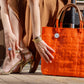 Woman wearing brown dress and gold heels touching orange woven recycled handbag in a city setting